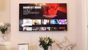 En väggmonterad tv-skärm som visar filmen ”DARKEST HOUR”, tillsammans med en Netflix-logotyp, en inramad tavla, en vas med blommor och en stjärnformad dekoration på ett konsolbord i trä.