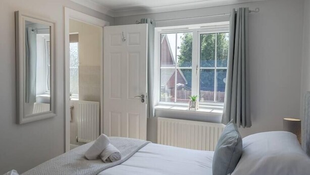 A bedroom with a bed, a window with curtains, a radiator, and a door leading to another room.