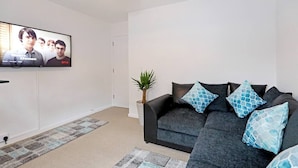 A modern living room with a large flat-screen TV mounted on the wall, a dark grey sofa with patterned cushions, a small potted plant, and a patterned rug. - "Stafford Rest" – Cozy Apartment with Smart TV, WiFi, and Stunning Street Views (Norfolk)