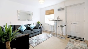 A modern living room with a grey sofa, a coffee table, and a large mirror. There is a window with blinds, a door, and a small bar area with stools. - "Stafford Rest" – Cozy Apartment with Smart TV, WiFi, and Stunning Street Views (Norfolk)