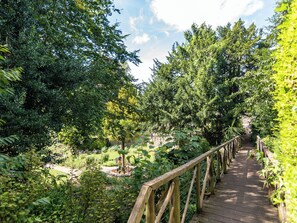 Una passerella in legno attraversa un giardino rigoglioso, con vegetazione fitta e un muro di pietra sullo sfondo.