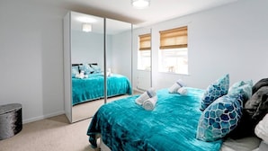 A bedroom with a large mirror, teal bedding, and a view of the outside through a window with blinds. - "Stafford Rest" – Cozy Apartment with Smart TV, WiFi, and Stunning Street Views (Norfolk)