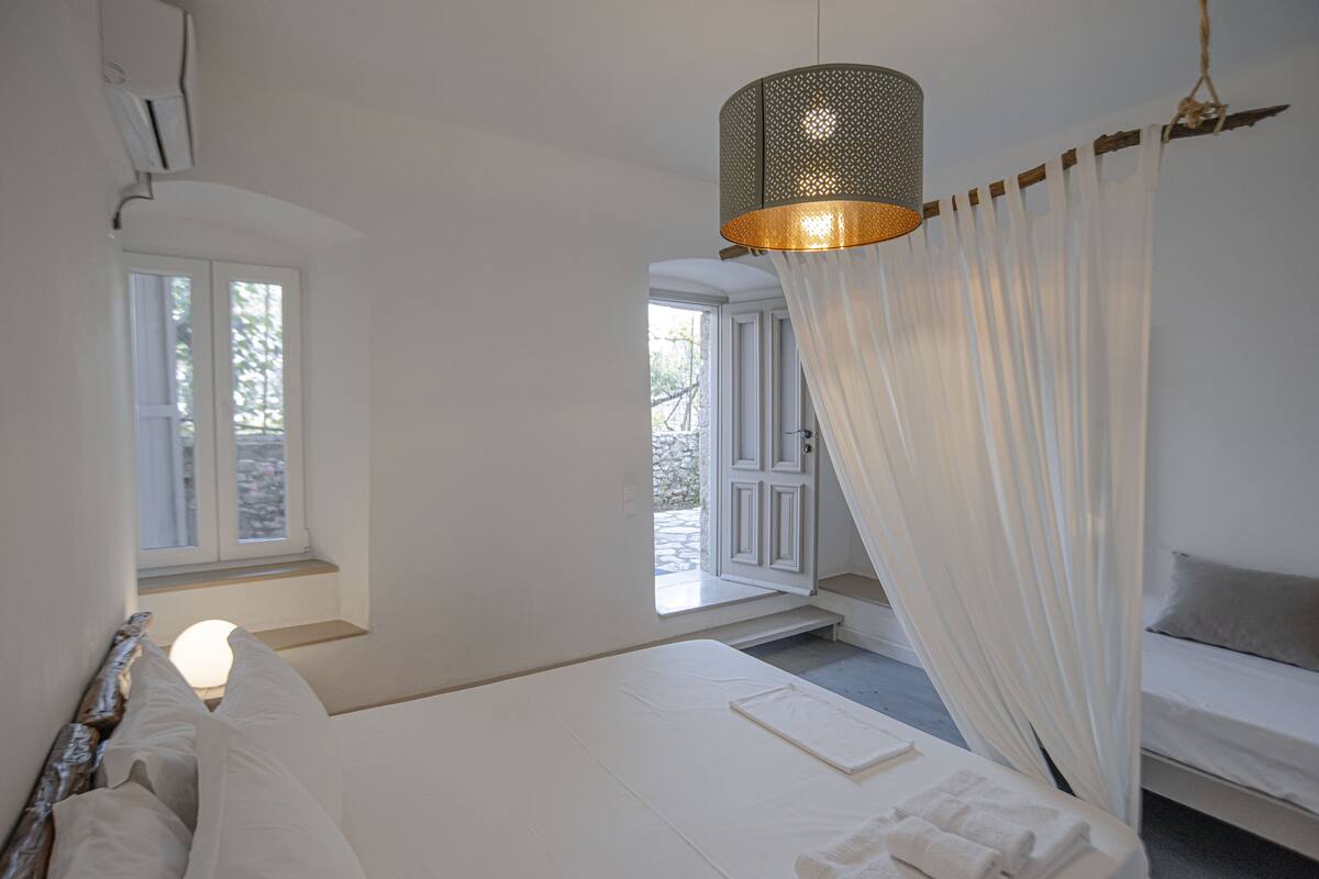 A bedroom with a bed, a bedside table, a hanging light fixture, and a view of the outside through a window.