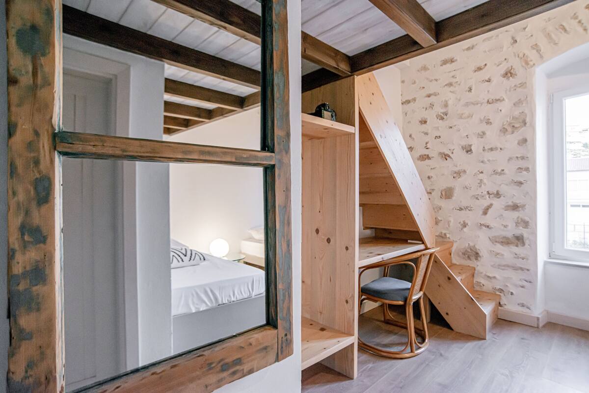 A room with a bed, a desk, and a chair. There is a staircase leading to an upper level.