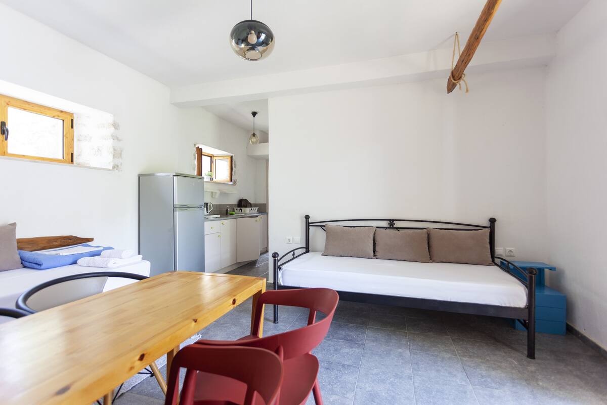 A compact room with a bed, a small kitchenette, a dining table with chairs, and a hanging light fixture.
