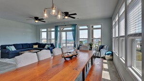 A spacious living room with a wooden dining table, blue sofa, and a view of the ocean through large windows. - Knot Salty: Ocean-view 4-bedroom for up to 11 in North Topsail Beach. (North Topsail Beach)