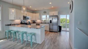A modern kitchen with white cabinets, a stainless steel refrigerator, and a large island with bar stools. - Knot Salty: Ocean-view 4-bedroom for up to 11 in North Topsail Beach. (North Topsail Beach)