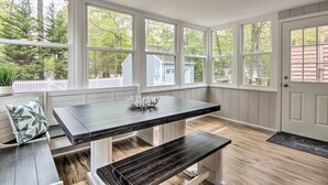 Una cucina con un tavolo e una panca in legno scuro, pareti bianche e ampie finestre che danno su alberi e un recinto bianco.