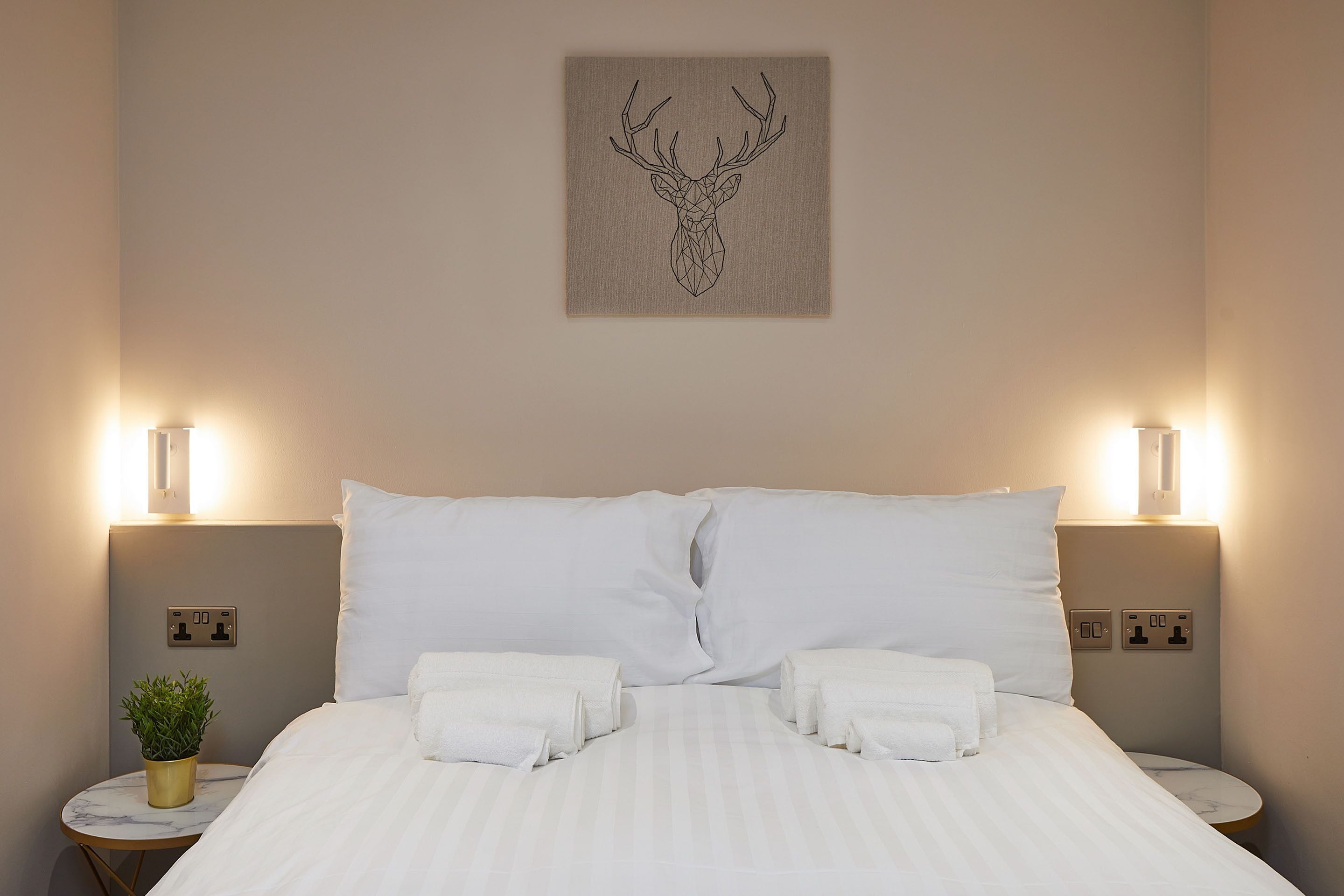 Ein ordentlich gemachtes Bett mit weißen Leinen, zwei Nachttischlampen und einem Wandbild mit einem Hirschkopf.
