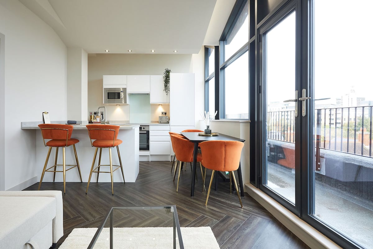 A modern kitchen with white cabinets, a stainless steel microwave, and a dining area with orange chairs and a black table.
