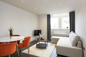 A modern living room with a round table, orange chairs, a sofa, and a TV on the wall.