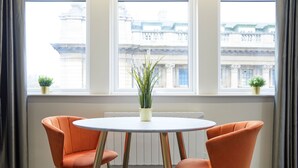 Une pièce avec une table ronde, deux chaises orange, une plante en pot et une vue sur les bâtiments à travers de grandes fenêtres.