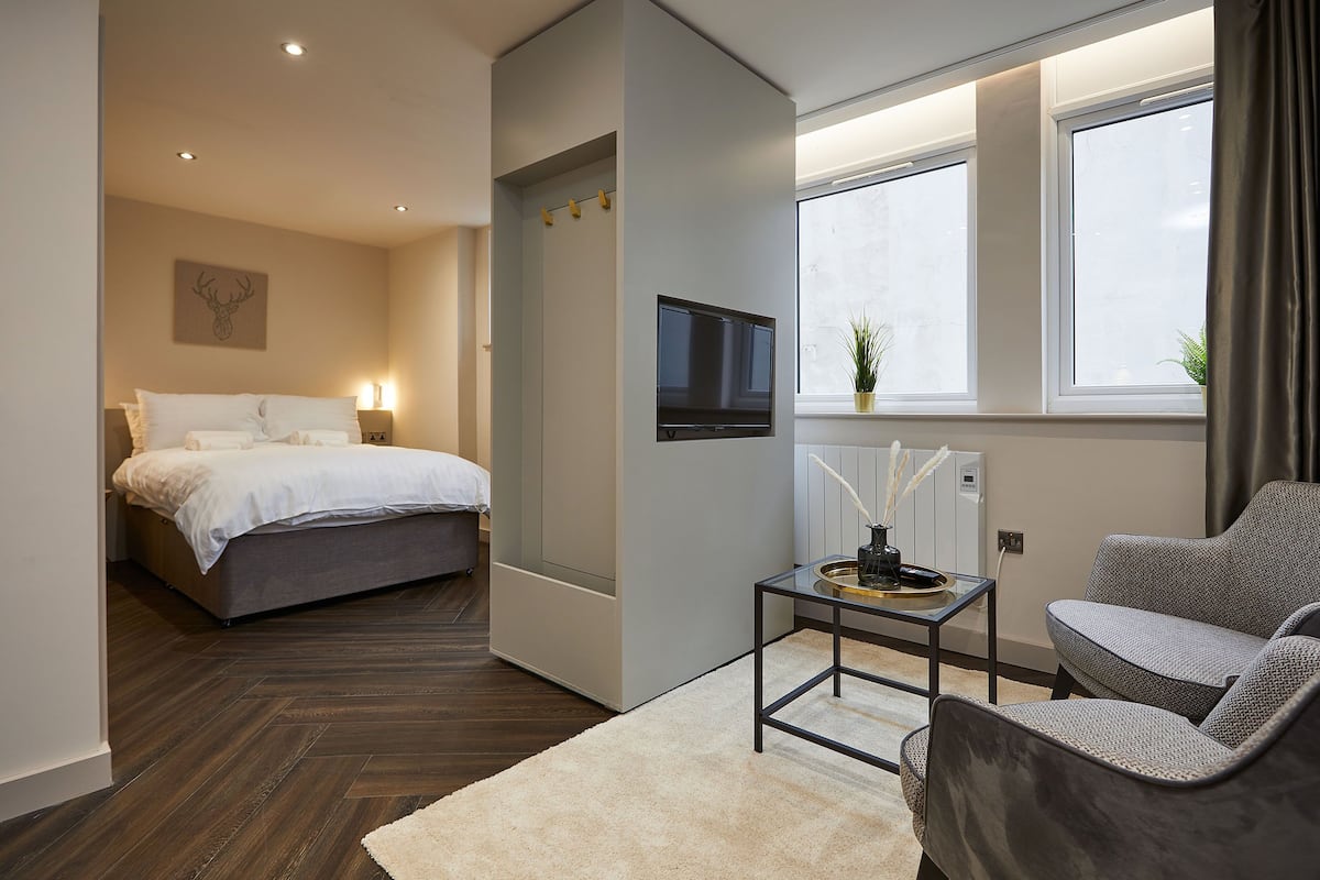 A modern hotel room with a bed, a TV, a seating area with a table, and large windows.