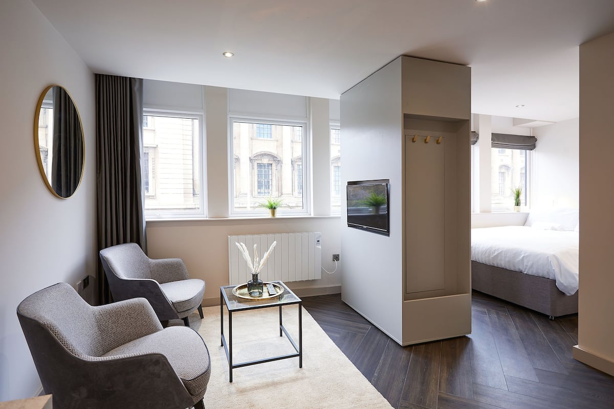 A modern hotel room with a bed, two armchairs, a small table, a TV, and large windows.