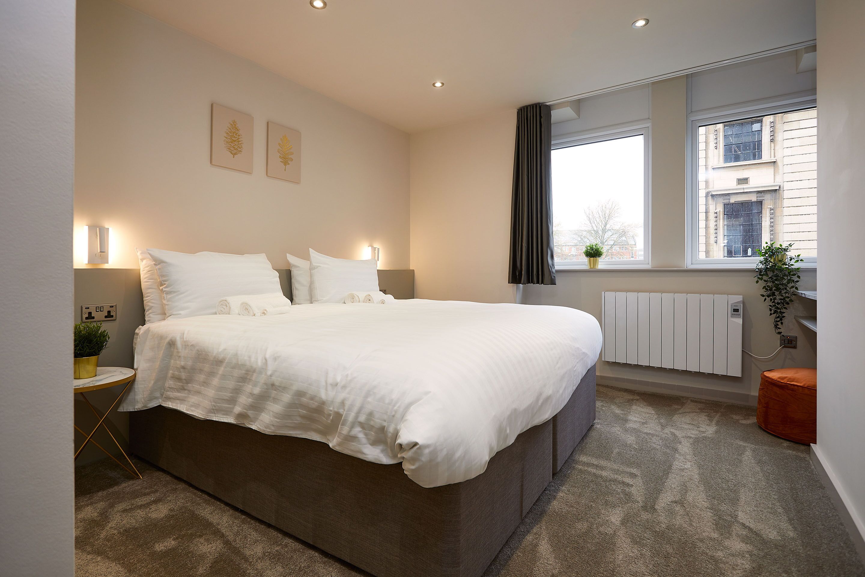 A modern bedroom with a large bed, bedside tables, a radiator, a window with curtains, and a view of buildings outside.