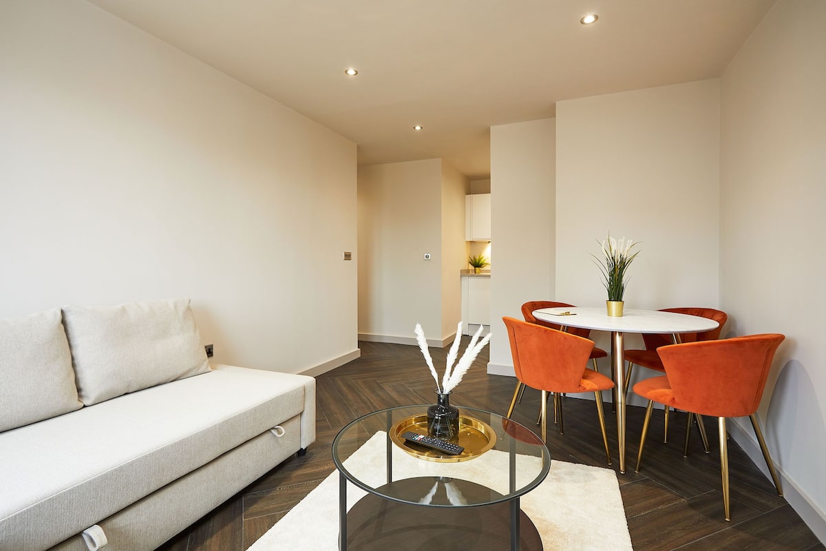A modern living room with a sofa, a glass coffee table, and a dining area with orange chairs and a white table.