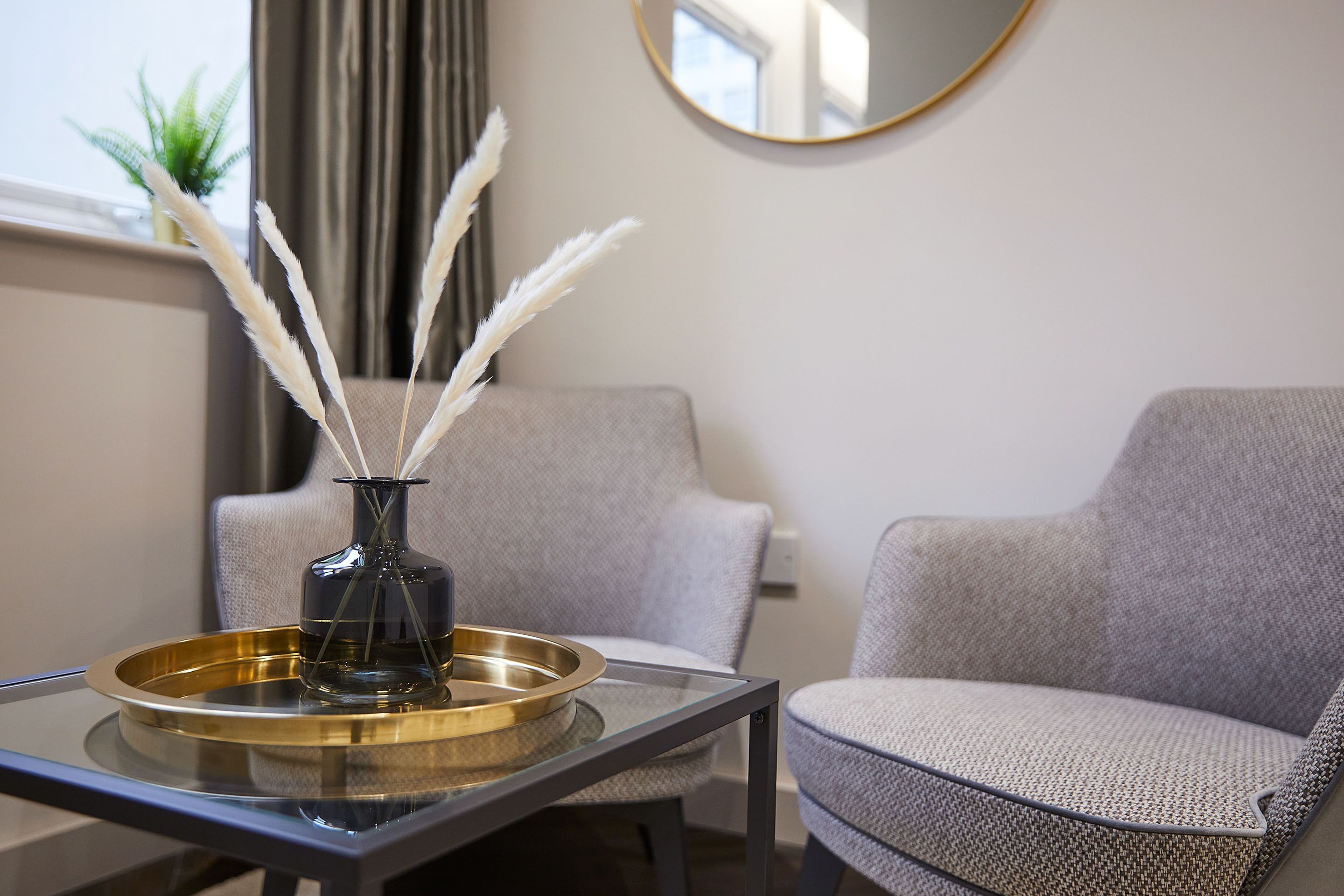 Un salon moderne avec une table basse en verre, un vase décoré de plumes blanches et deux fauteuils gris.