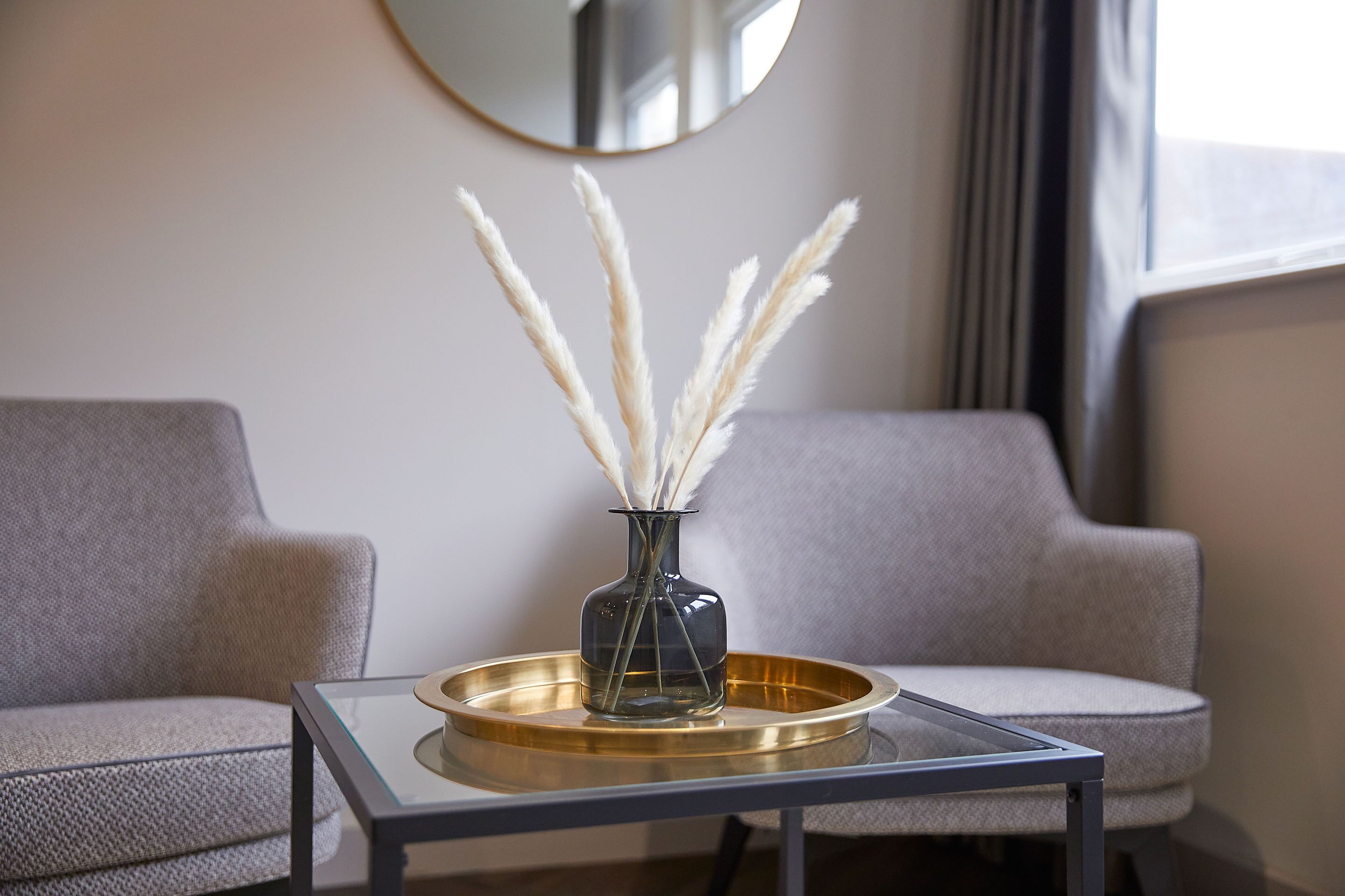 Un salon moderne avec une table basse en verre, un vase contenant de l’herbe des pampas et deux canapés gris.