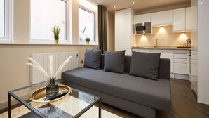 Un salon moderne avec un canapé gris, une table basse en verre et, au fond, une vue sur la cuisine.