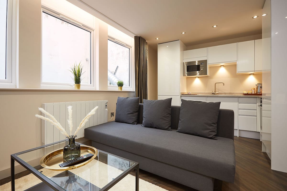 A modern living room with a grey sofa, a glass coffee table, and a view of the kitchen in the background.