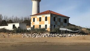 Un bâtiment situé sur la côte, avec un phare en arrière-plan, et qui se trouve sur une plage de sable parsemée de rochers.