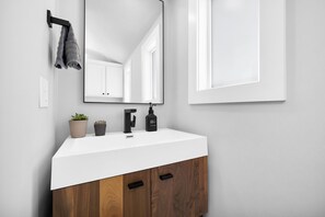 A bathroom with a white sink, a mirror, a towel rack, two potted plants, and a wooden cabinet with two drawers. - Roxie's Modern Lake Front House on the water! (Russells Point)