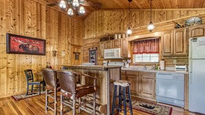 Une cabane en bois rustique dotée d’une cuisine avec un îlot central, des tabourets de bar, un ventilateur de plafond et un tableau représentant un élan accroché au mur.