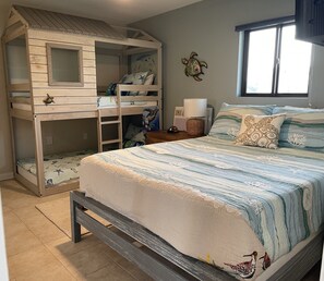 Un dormitorio con literas, una cama con colcha a rayas, una mesita de noche con lámpara y una decoración de pared con forma de tortuga.