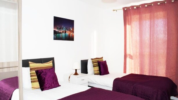 A hotel room with two beds, a nightstand, a lamp, a cityscape picture on the wall, and a red curtain. - TMS Beautiful 2BR Apartment! Ensuite! Lakeside (Grays)