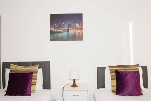 A hotel room with two beds, a nightstand with a lamp, and a cityscape picture on the wall.