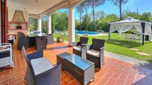 An outdoor patio with wicker furniture, a brick fireplace, and a view of a swimming pool and trees. - Villa Cerbaie (Fucecchio)