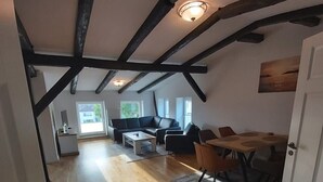 A spacious loft with exposed wooden beams, a dining area with a wooden table and chairs, a sofa, and a view of the outside. - House (Klein Upahl)