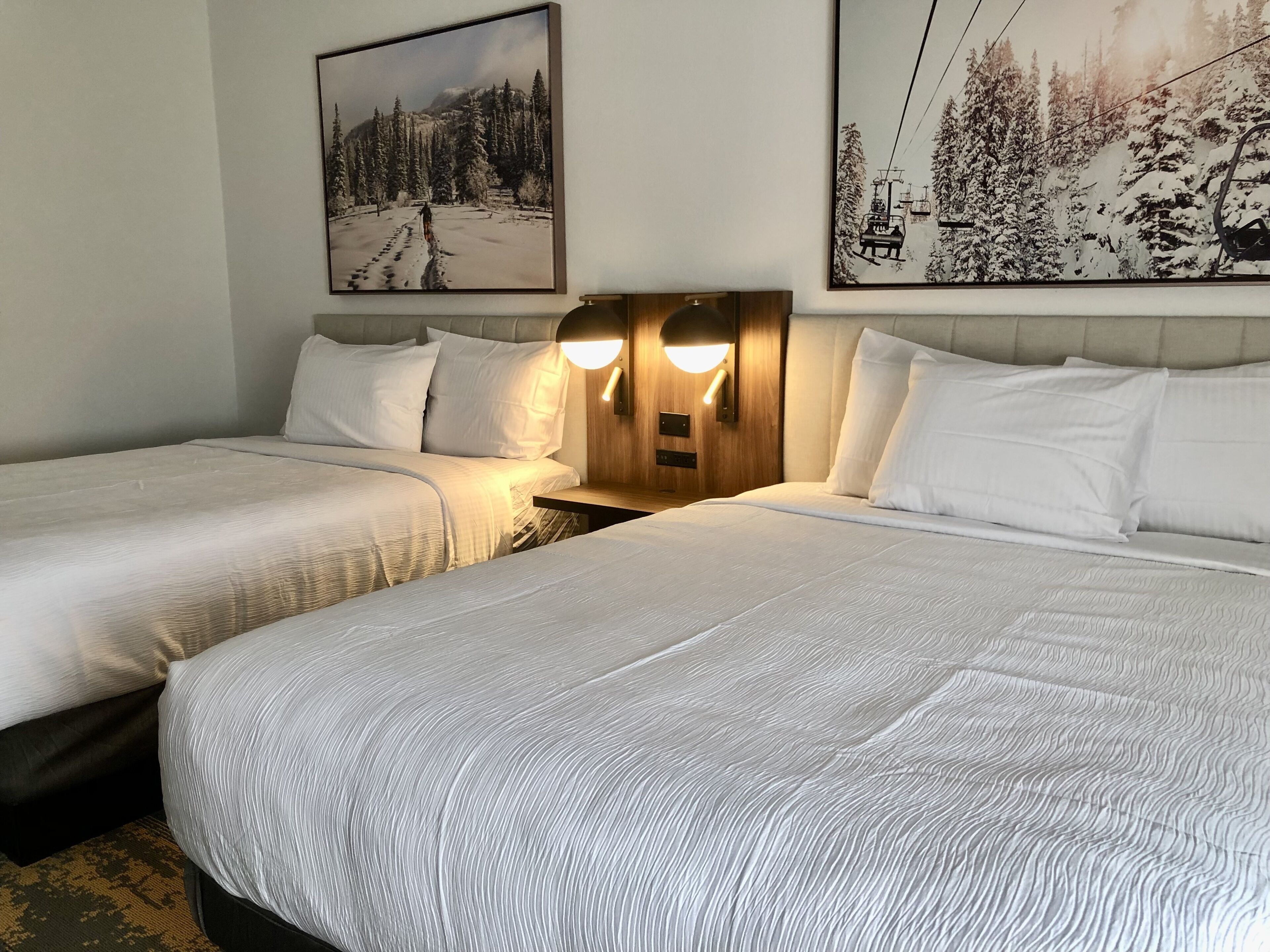 A hotel room with two beds, a wooden headboard with wall-mounted lights, and two framed pictures depicting snowy landscapes.