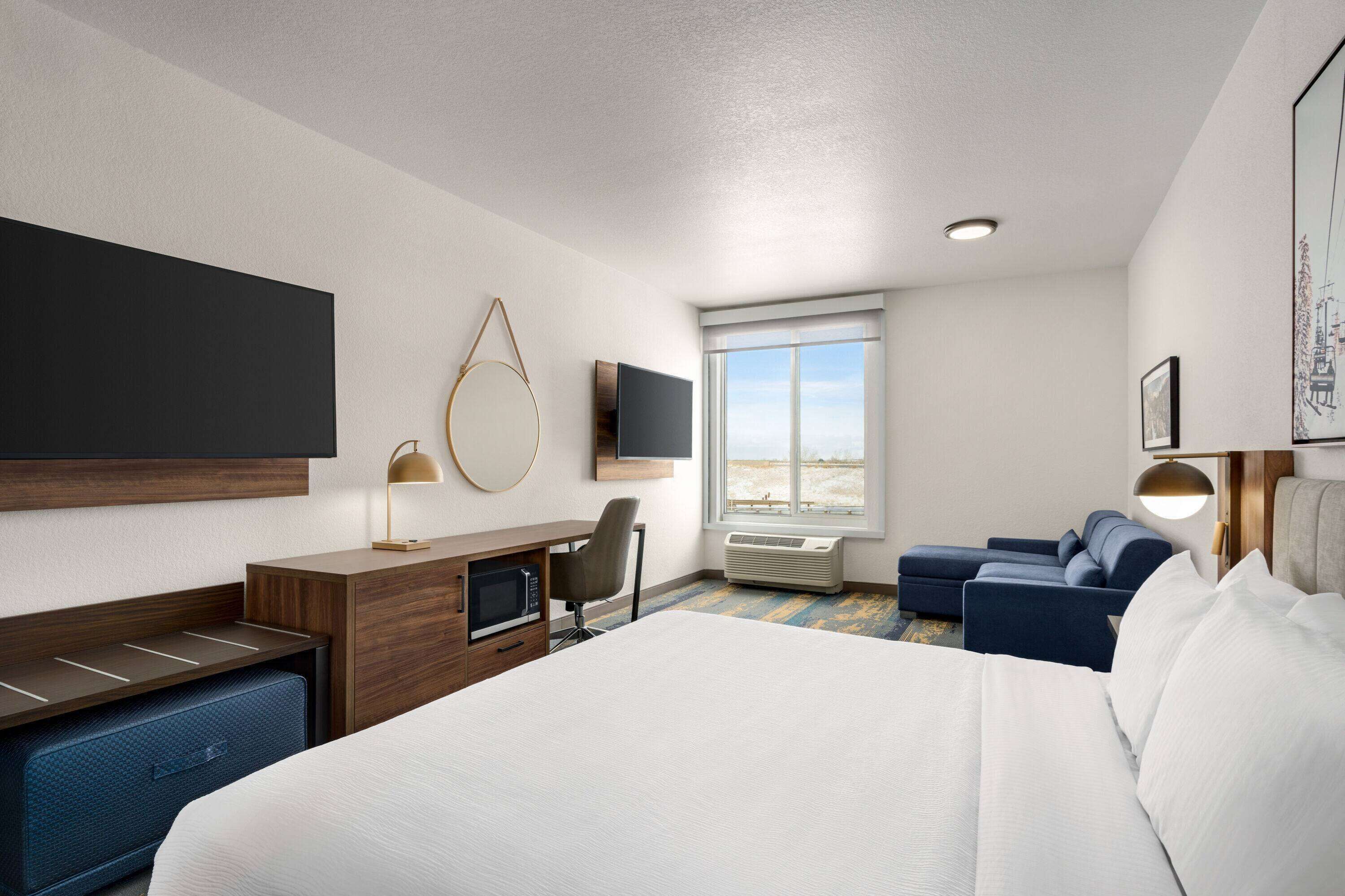 A hotel room with a large bed, a desk with a computer, a chair, a sofa, a television, and a window with a view.