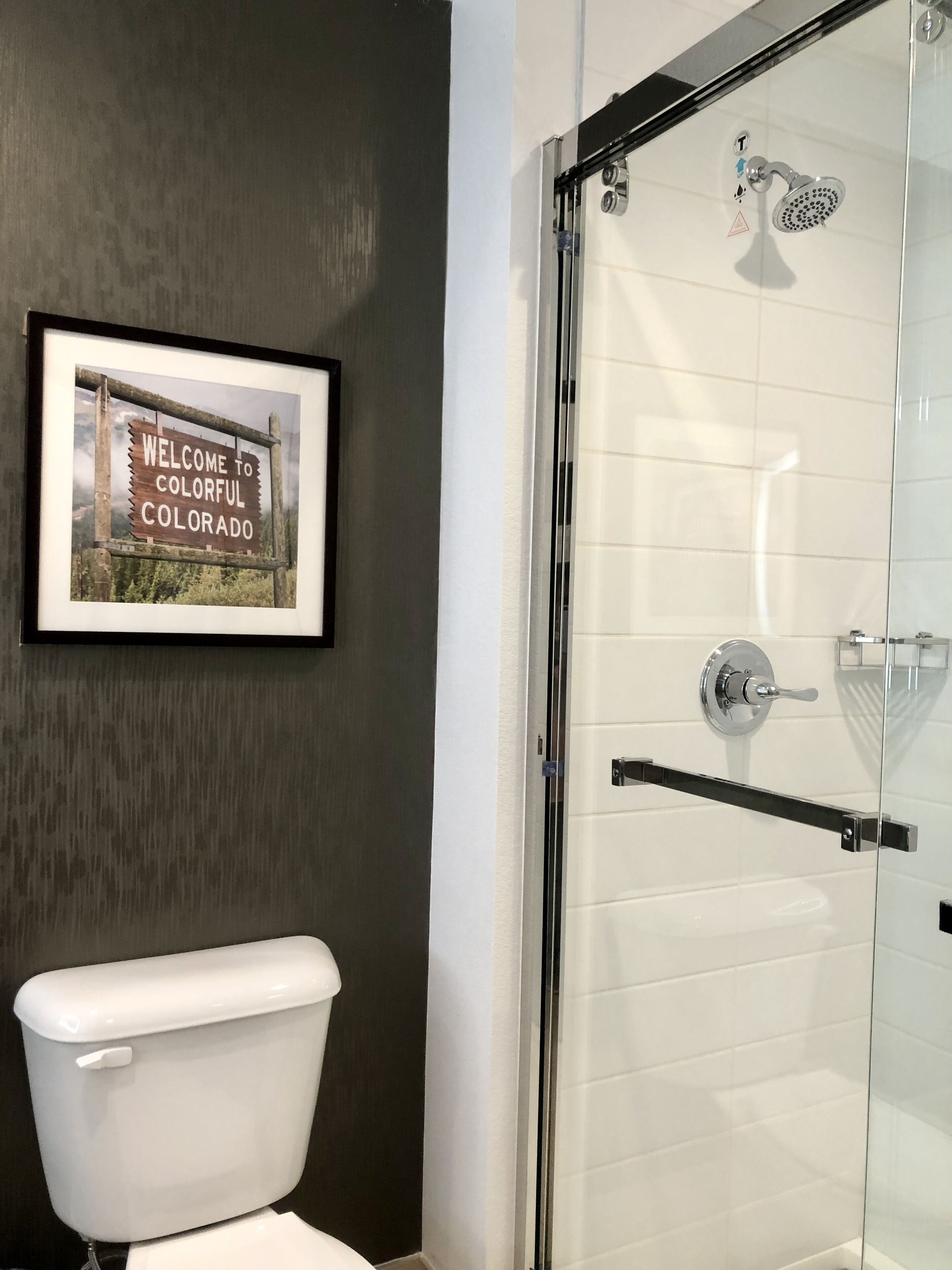 A bathroom with a white toilet, a framed welcome sign, and a glass-enclosed shower.