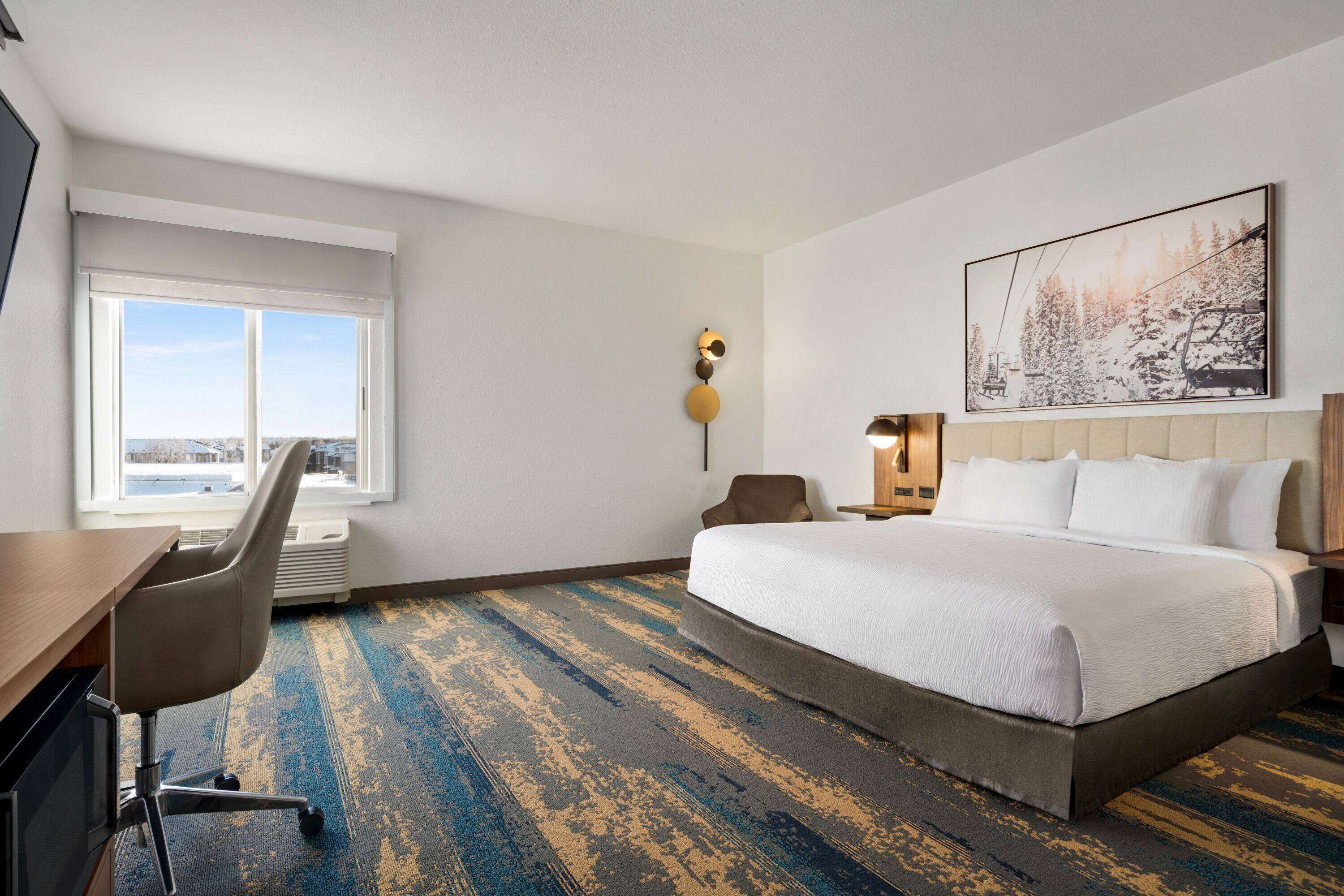 A hotel room with a large bed, a desk with a chair, a window with a view, and a wall-mounted artwork.