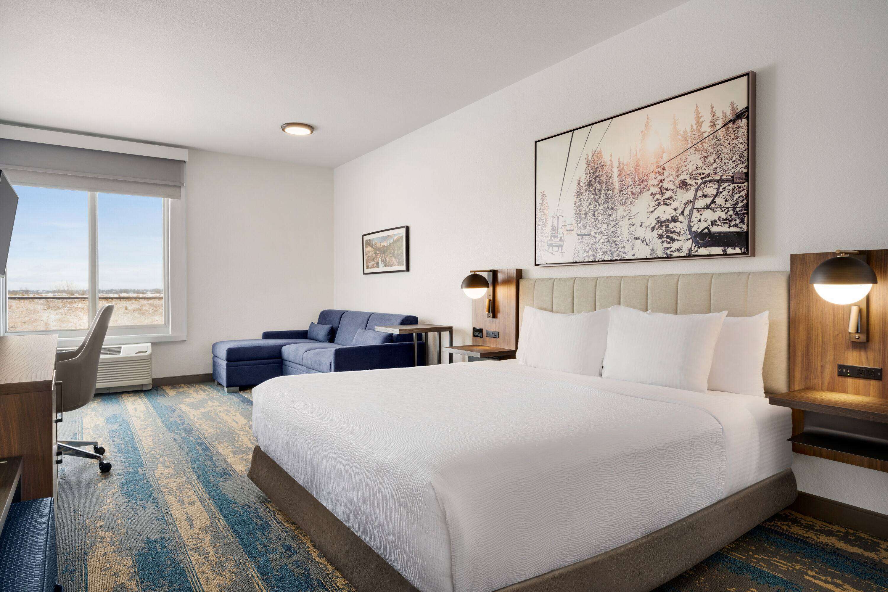 A hotel room with a large bed, a desk, a chair, a sofa, a window with a view, and a painting on the wall.
