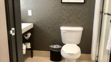 A bathroom with a toilet, a trash bin, a sink, and a framed welcome sign.