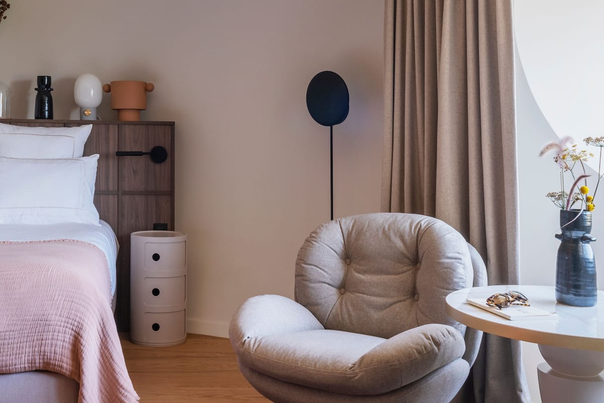 A bedroom with a bed, a chair, a bedside table, a lamp, and a vase with flowers.