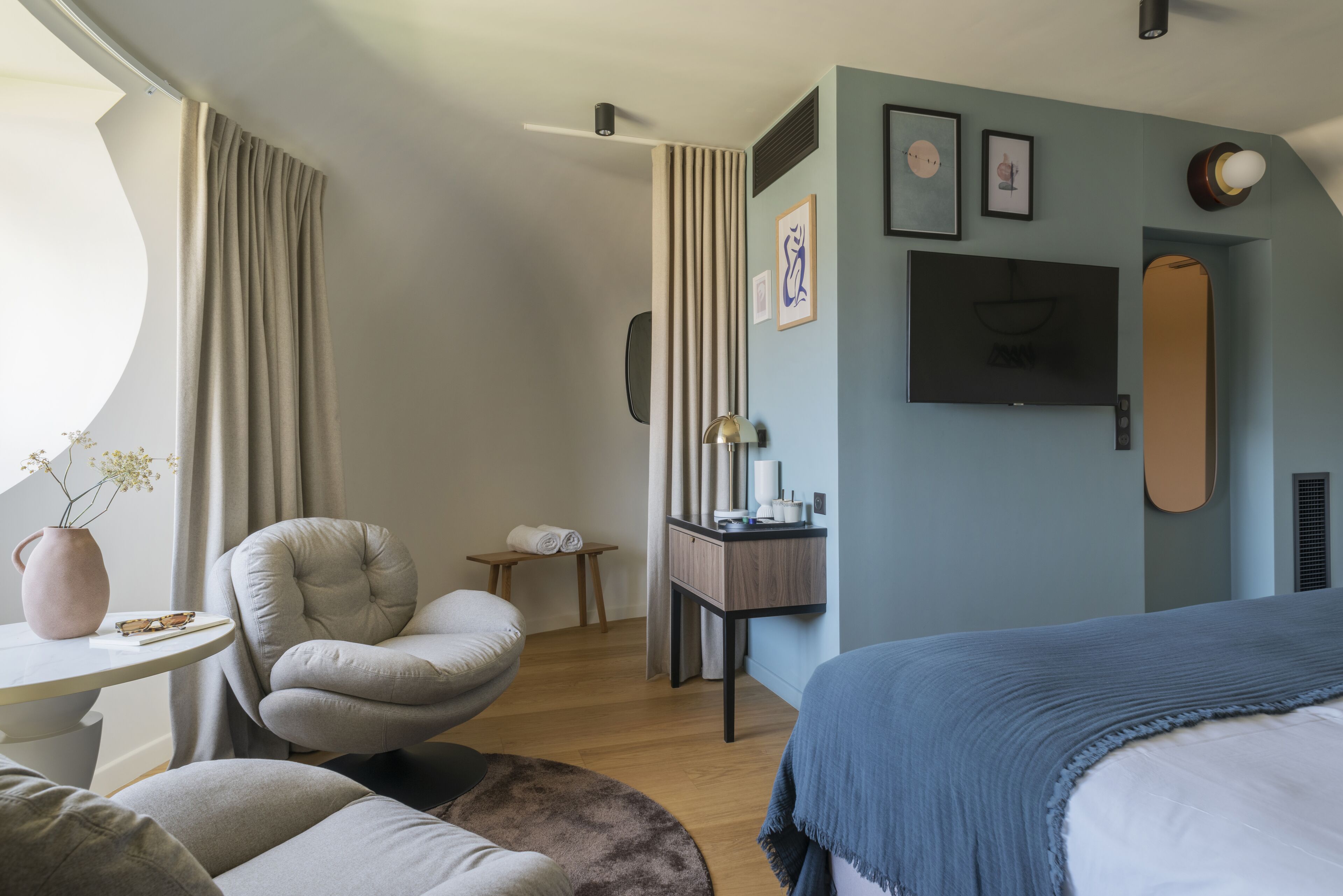 A modern hotel room with a bed, a seating area with a chair, a small table with a vase, and a TV mounted on the wall.