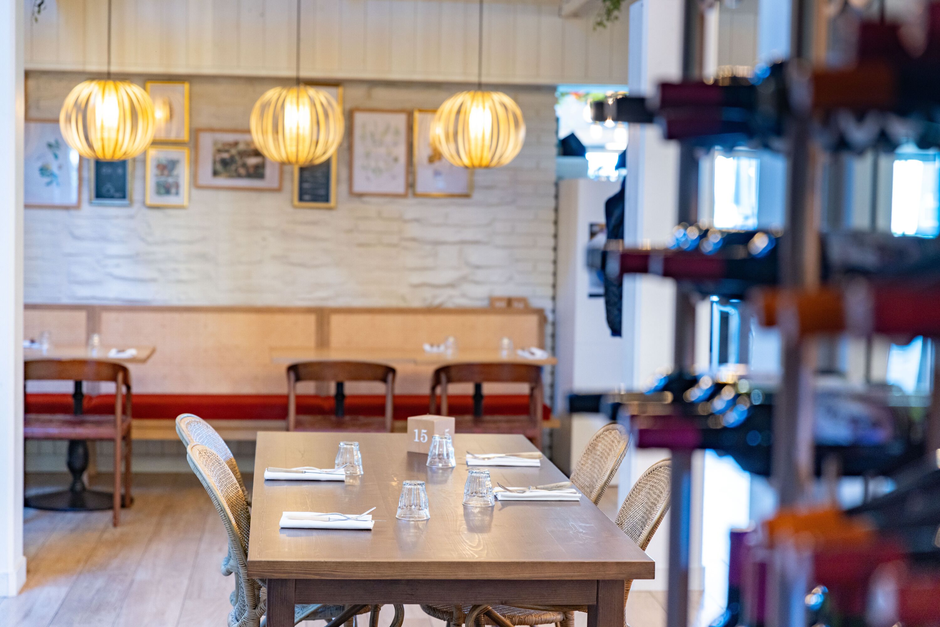 Ein Restaurant mit Holztischen und -stühlen, hängenden Pendelleuchten, gerahmten Bildern an der Wand und einem Weinkühlschrank im Hintergrund.