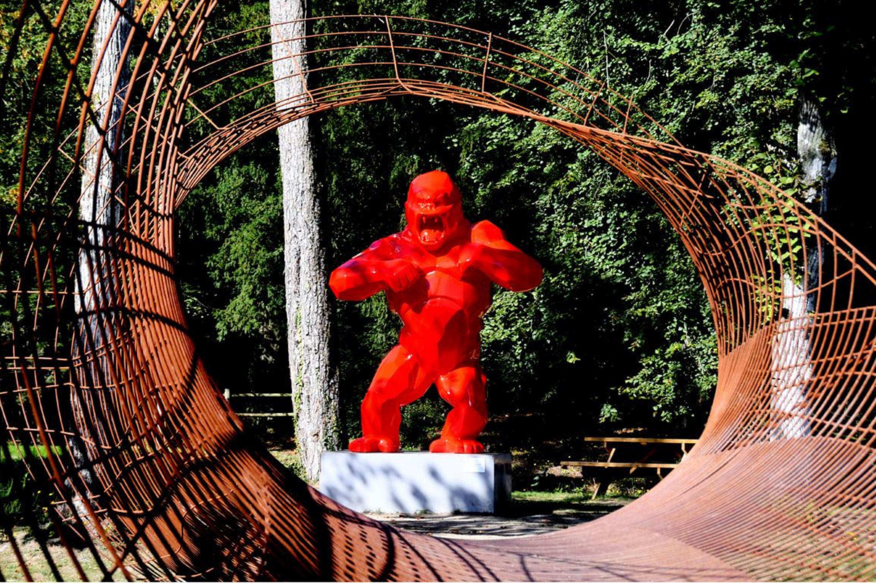 Estatua roja de una figura musculosa en una pose dinámica, rodeada por una estructura de arco de metal en un parque.