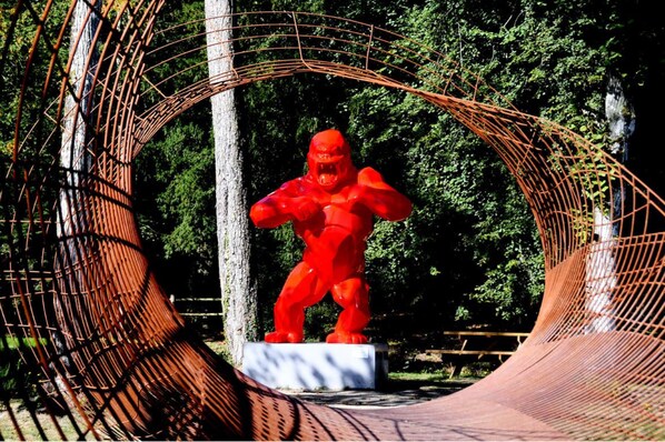 Estatua roja de una figura musculosa en una pose dinámica, rodeada por una estructura de arco de metal en un parque.