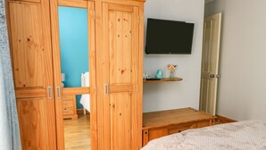 A bedroom with a wooden wardrobe, a bed, a TV mounted on the wall, and a small shelf with decorative items. - Aurora (Holyhead)