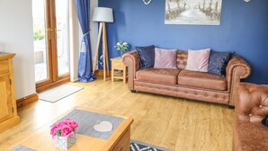 A living room with a brown leather sofa, a wooden coffee table with pink flowers, a lamp, and a painting of a beach scene on the wall. - Aurora (Holyhead)