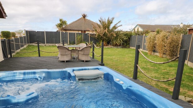 A hot tub with seating and a table, surrounded by a fenced yard with grass and plants. - Aurora (Holyhead)