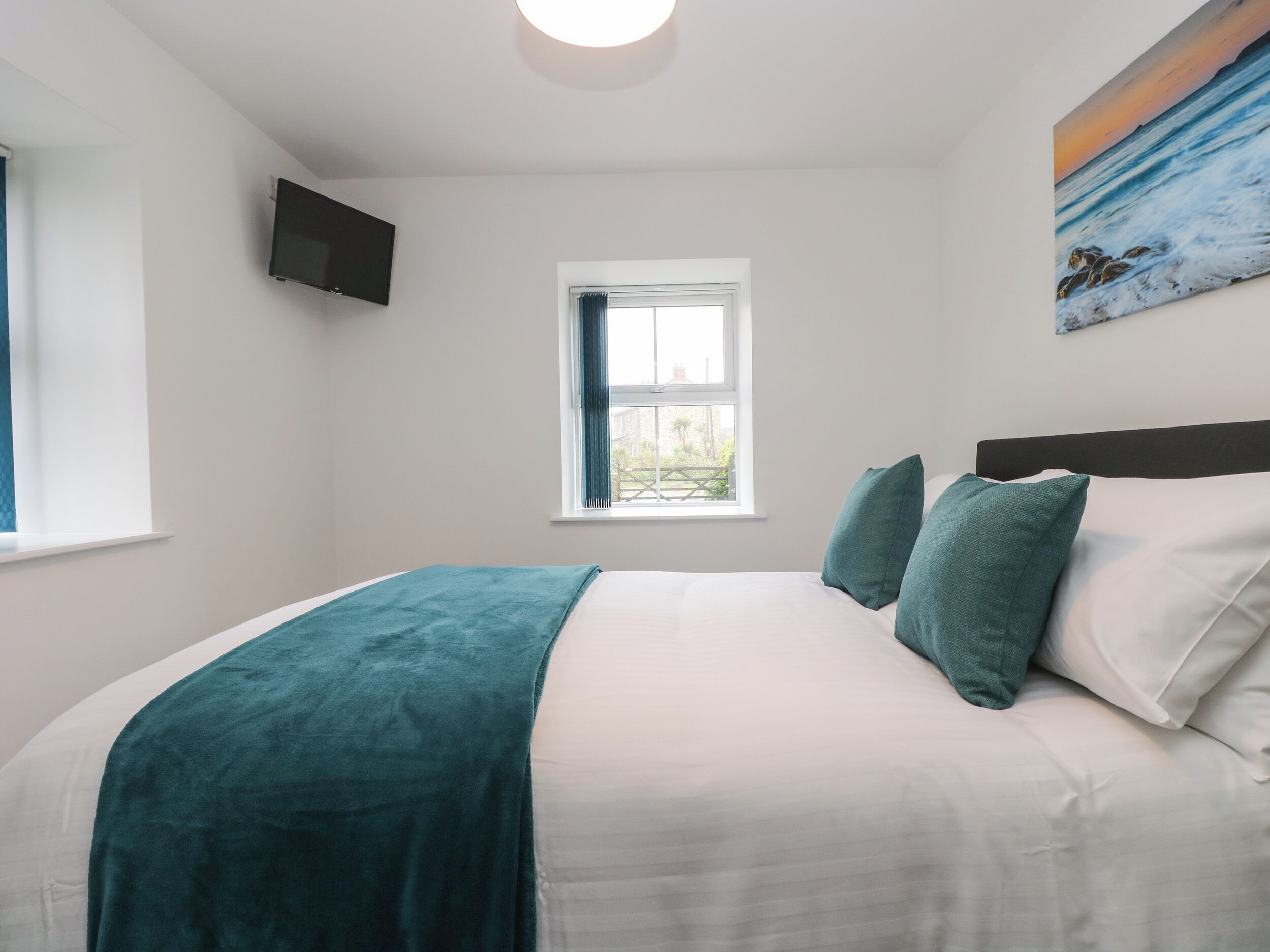 Un dormitorio con cama, ventana, un cuadro y televisión en la pared.