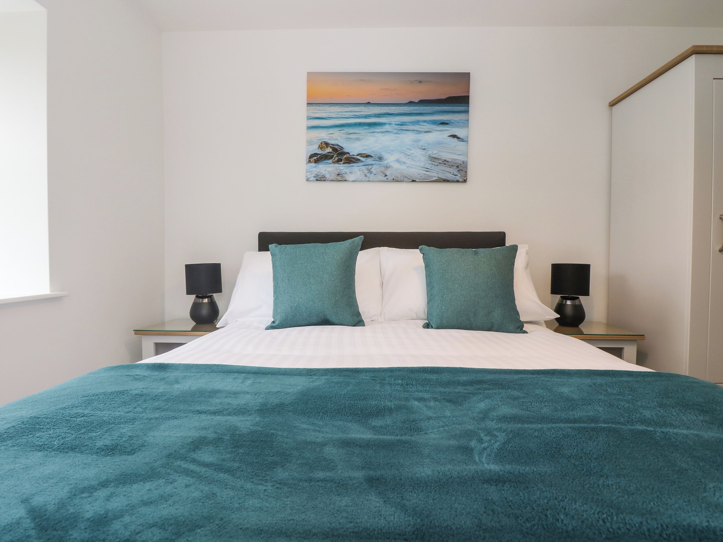 A bedroom with a bed, two nightstands, a painting of a beach scene, and a teal throw blanket.