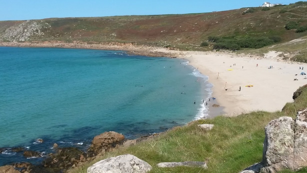 Une plage côtière aux eaux turquoise et limpides, bordée d’un rivage sablonneux et d’une colline herbeuse, avec un bâtiment visible au loin.
