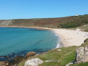 A coastal beach with clear blue waters, sandy shore, and a grassy hill with a building in the distance. - Wellfield Cottage (Penzance)